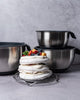 Stack of meringue dessert with fruit on a metal stand, surrounded by metallic cookware on a gray background
